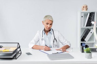 Gri saçlı kıdemli doktor masasının yanına reçete yazıyor. 