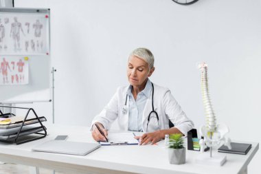 Gri saçlı doktor masasının yanına reçete yazıyor. 