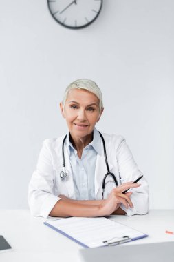 Beyaz önlüklü mutlu kıdemli doktor masasının yanında elinde kalem tutuyor. 