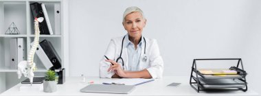 gülümseyen doktor kalemini masanın yanında, pankartta tutuyor