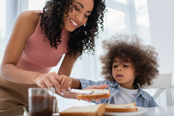 Smiling african american mother spreading chocolate paste on bread near kid at home 