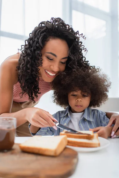 African american woman spreading chocolate paste on bread near kid