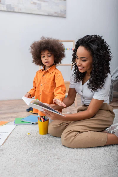 Mulher Africana Positiva Segurando Livro Perto Filha Lápis Cor Casa — Fotografia de Stock