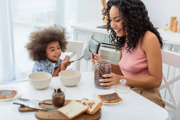 幸せなアフリカ系アメリカ人の母親と子供の近くに座ってシリアルとパンとチョコレートペーストでキッチン — ストック写真