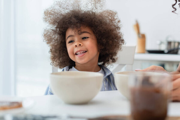 Smiling african american kid sitting near bowls and mom in kitchen 