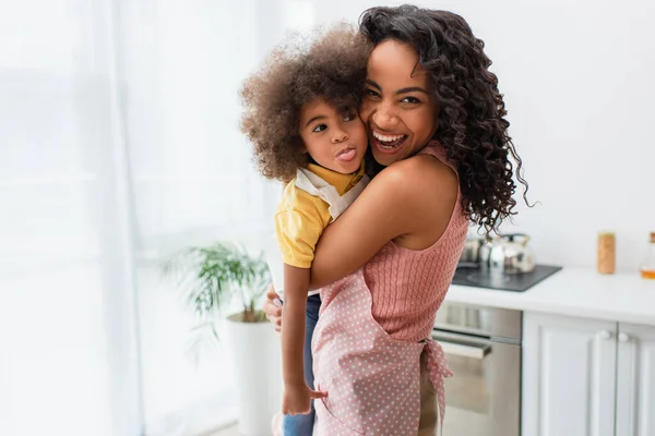 Šťastná Africká Americká Matka Zástěře Objímající Dítě Vyčnívající Jazyk Kuchyni — Stock fotografie