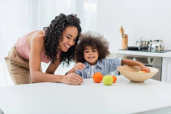 幸せなアフリカ系アメリカ人の母親はキッチンで果物と子供の近くに立って — ストック写真