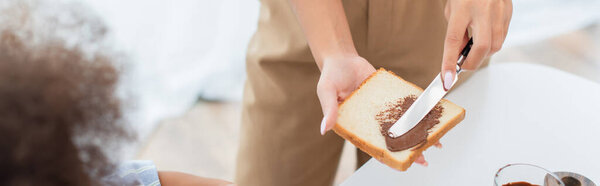 Обрезанный вид на африканскую американскую мать делает тост с шоколадной пастой рядом с ребенком, баннер 