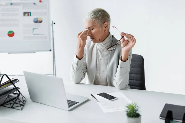 tired businesswoman with grey hair touching eyes and holding glasses near laptop