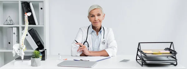 Sonriente Médico Sosteniendo Pluma Cerca Dispositivos Escritorio Pancarta — Foto de Stock