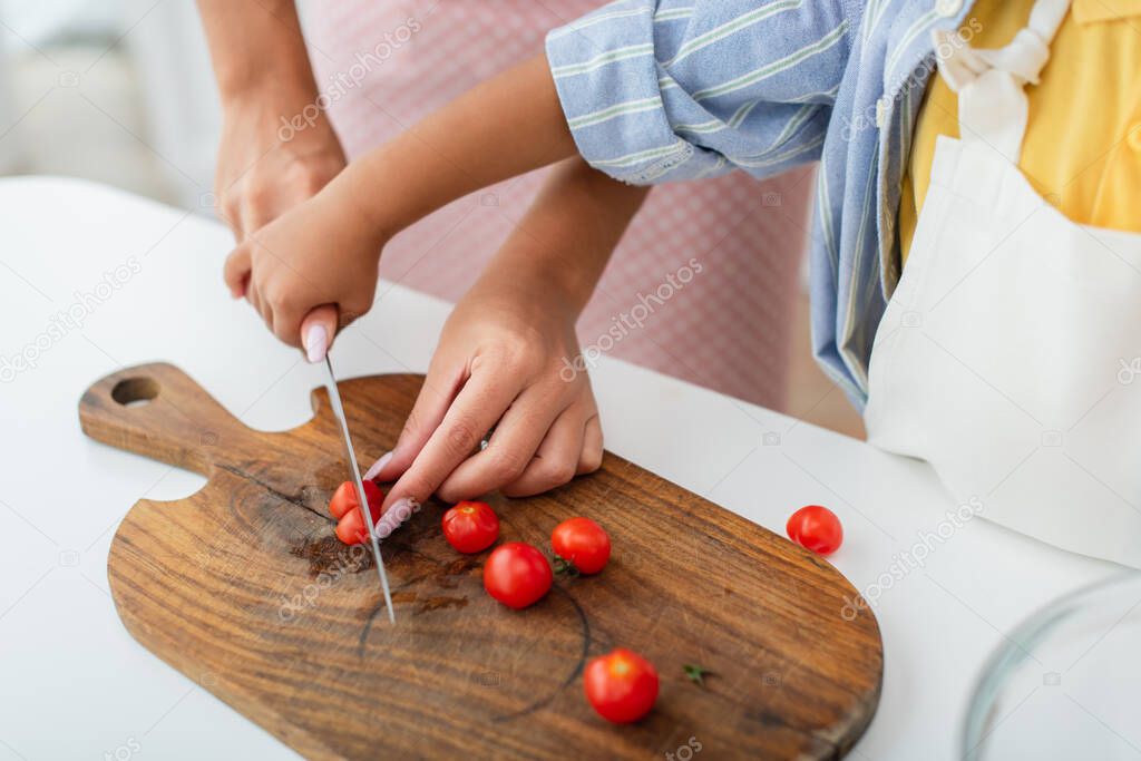 Cropped view of african american kid holding hand of mother with knife cutting cherry tomatoes in kitchen 