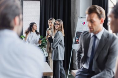 happy woman holding coffee to go near colleagues and blurred multiethnic businessmen clipart