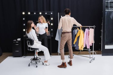 african american stylist choosing clothes while makeup artist doing visage to model in dressing room clipart