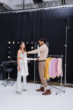 full length view of african american stylist dressing young brunette woman in photo studio clipart