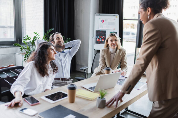 young african american businessman talking to excited colleagues laughing at desk in office