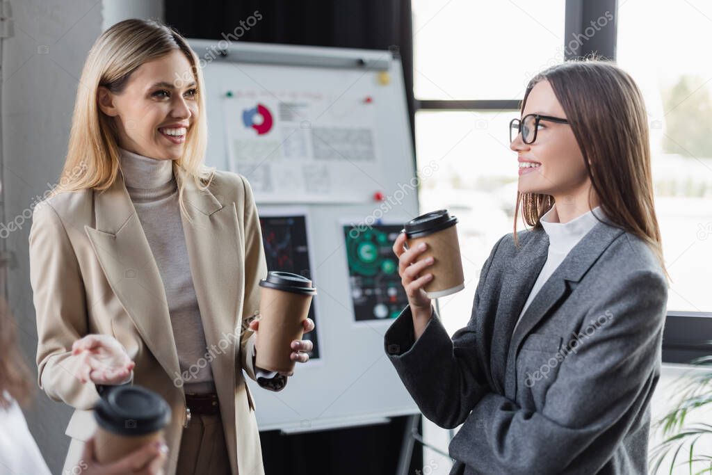 cheerful businesswomen with paper cups talking near blurred flip chart in office