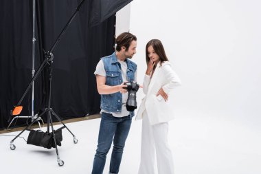 Fotoğrafçı, fotoğraf stüdyosunda eli ağızlı, şaşırmış bir modele dijital kamera gösteriyor.