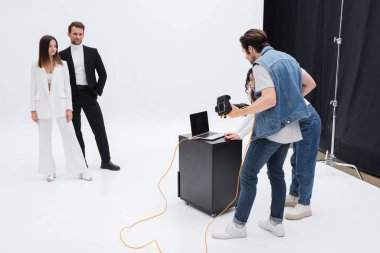 art director with photographer looking at laptop with blank screen near models in photo studio on white clipart