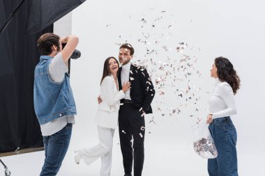 cheerful models in suits posing during photo session near assistant throwing confetti clipart