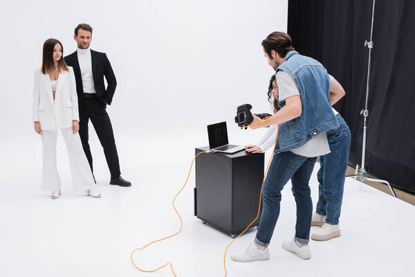 art director with photographer looking at laptop with blank screen near models in photo studio on white