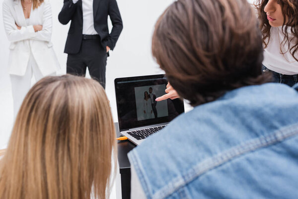 art director pointing at laptop monitor near blurred colleagues and stylish models isolated on white
