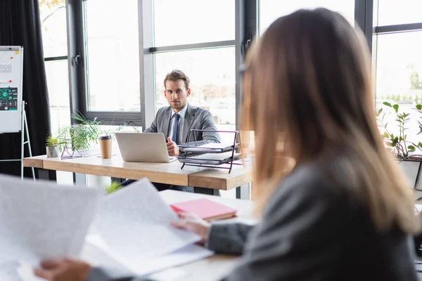 Biznesmen Patrząc Zamazanego Kolegę Podczas Siedzenia Przy Biurku Pobliżu Laptopa — Zdjęcie stockowe