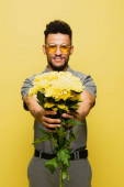 rozmazané africký Američan muž v slunečních brýlích a šedé tenisové košili drží kytice květin izolovaných na žluté 