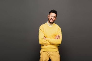 Sarı elbiseli ve gözlüklü neşeli Afro-Amerikan adam koyu gri üzerinde çapraz kollarıyla ayakta duruyor.