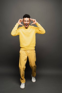 amazed african american man in glasses and yellow outfit smiling on dark grey clipart