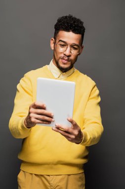 Genç Afro-Amerikan gözlüklü adam gri üzerinde izole edilmiş dijital tablet kullanıyor.