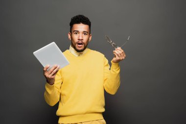 Koyu gri üzerinde gözlük ve dijital tablet tutan Afro-Amerikan adam şok oldu.