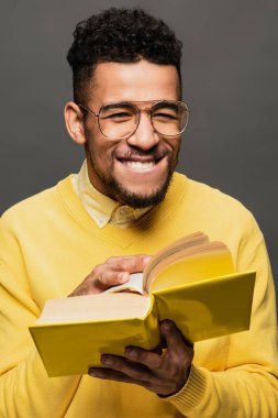 cheerful african american man in glasses and yellow sweater holding book isolated on dark grey clipart
