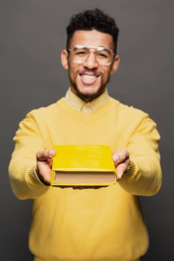 Gözlüklü ve sarı elbiseli Afro-Amerikan adam koyu gri üzerine kitap tutuyor.