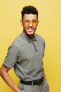 happy african american man in sunglasses and grey tennis shirt standing with hands in pockets isolated on yellow  clipart