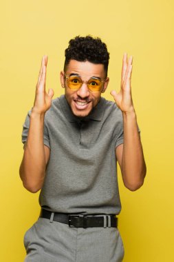 smiling african american man in sunglasses and grey tennis shirt gesturing isolated on yellow clipart