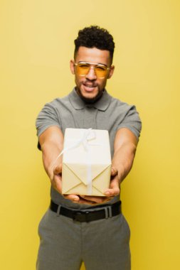 Bulanık Afro-Amerikalı güneş gözlüklü ve gri tenis tişörtlü adam sarıya sarılmış hediyesini tutuyor.