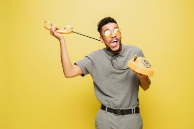 Güneş gözlüklü Afro-Amerikan adam kendini telefon kablosuyla sarıya boğarken dilini çıkartıyor. 