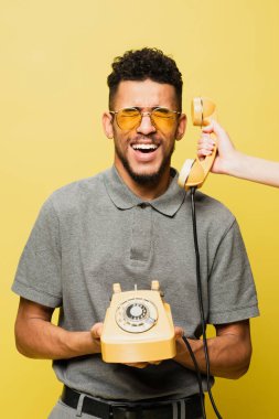 Güneş gözlüklü, sarı tişörtlü, Afro-Amerikan bir adamın yanında cep telefonu tutan kadın. 
