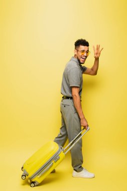 Güneş gözlüklü, gri tenis tişörtlü, bavullu ve sarı eli sallayan mutlu Afro-Amerikan bir adam.