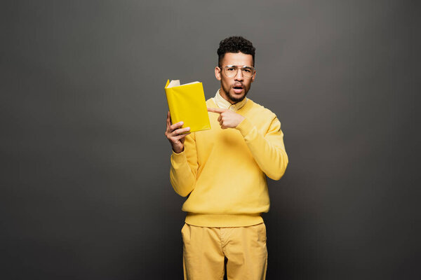 surprised african american man in glasses and yellow outfit pointing at book on dark grey
