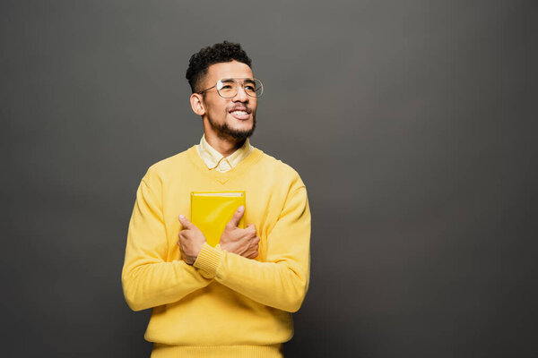 smiling african american man in glasses and yellow outfit holding book on dark grey