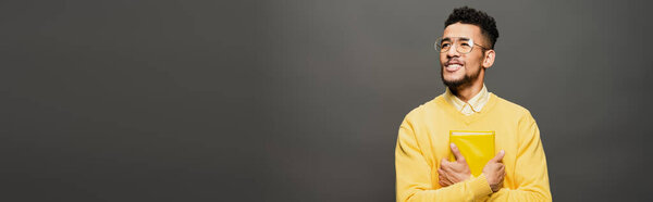 smiling african american man in glasses and yellow outfit holding book on dark grey, banner