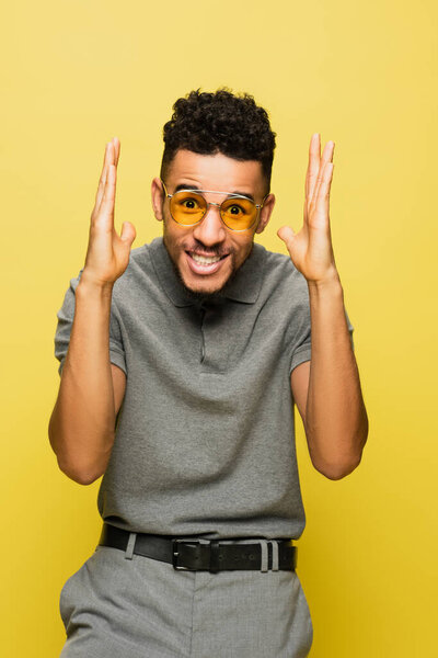 smiling african american man in sunglasses and grey tennis shirt gesturing isolated on yellow