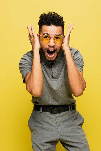 shocked african american man in sunglasses and grey tennis shirt gesturing isolated on yellow 
