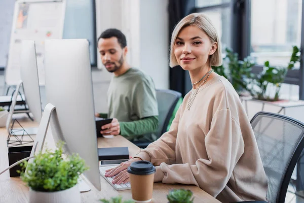 Blonde Woman Looking Camera Computer Monitor Blurred African American Colleague — Stockfoto