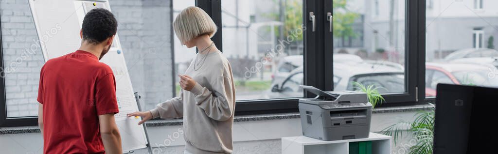 Interracial colleagues looking at flip chart in modern office, banner
