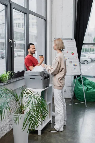 Sarışın kadın, elinde kağıt bardakla Afrikalı Amerikalı meslektaşının yanında evrak kopyalıyor. 