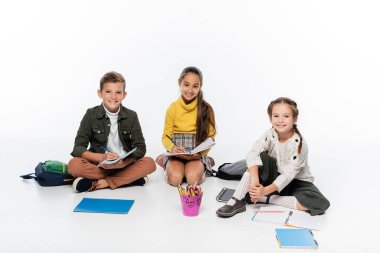 cheerful schoolboy and schoolgirls sitting near backpacks and notebooks while drawing on white  clipart