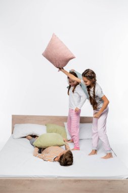 Pijamalı neşeli kızlar yastık savaşı yaparken beyaz tenli bir erkeğin yanında dikiliyorlar.
