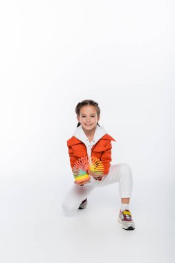 full length of cheerful kid in orange jacket playing with rainbow slinky on white clipart